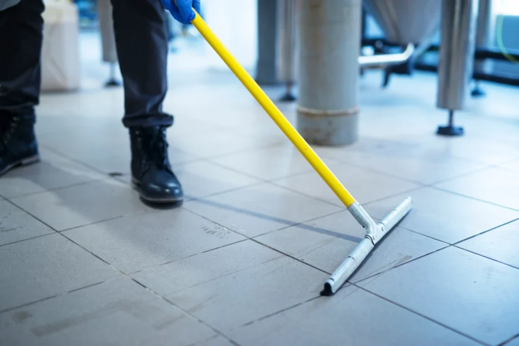 A person wearing dark boots and blue gloves using a large squeegee to push water across a light-colored tiled floor.