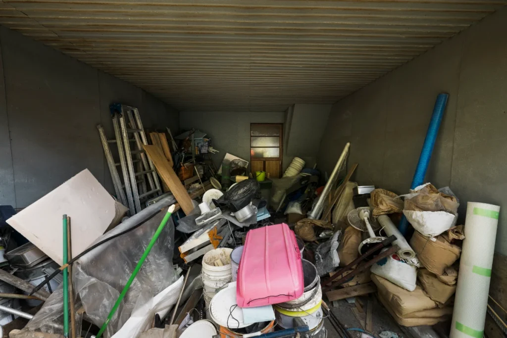 A heavily cluttered garage or storage space filled with debris, old equipment, and various discarded items, typical of a site needing foreclosure cleaning services.