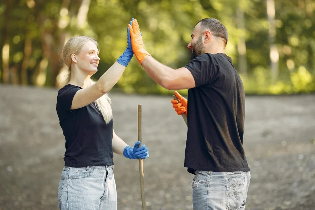 Two people wearing rubber gloves high-five each other while holding outdoor cleanup tools.