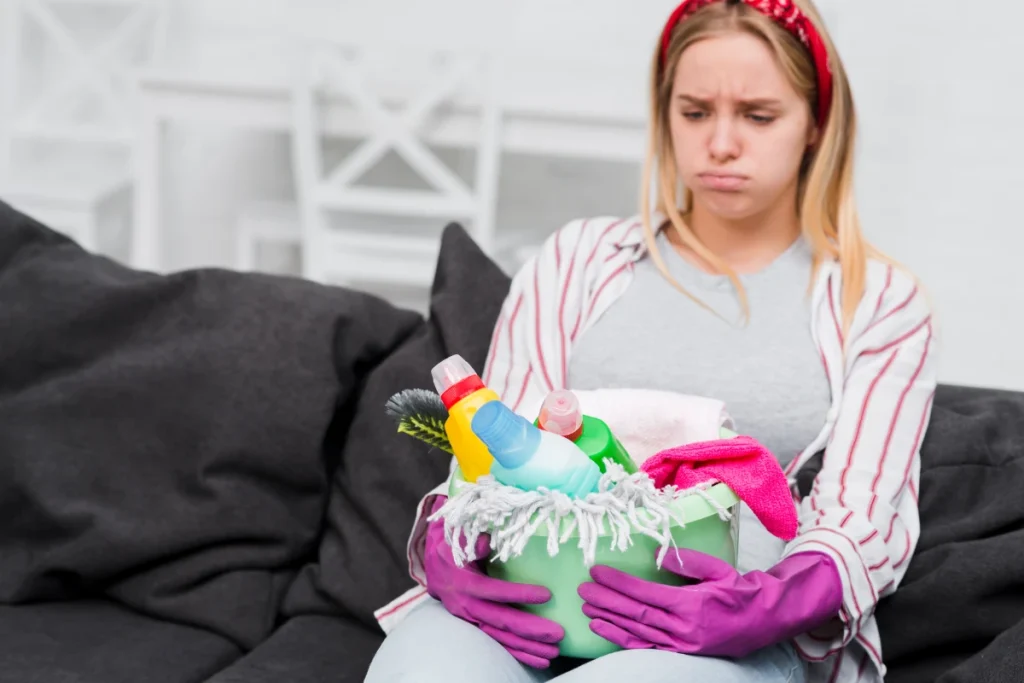 A frustrated person sitting on a couch holding a bucket overflowing with cleaning supplies, representing the mental load of housework.
